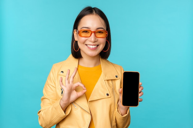 Mujer coreana sonriente que muestra la interfaz de la aplicación de teléfono móvil aplicación de teléfono inteligente recomendando en el teléfono celular de pie sobre fondo azul