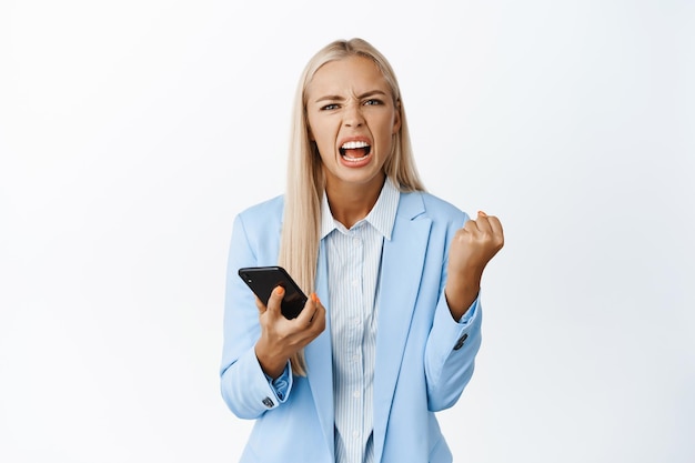 Mujer corporativa enojada y cabreada sosteniendo el puño del teléfono móvil y gritando de pie de forma agresiva en traje sobre fondo blanco