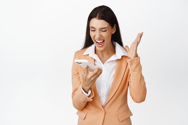 Mujer corporativa enojada gritando a su teléfono gritando en altavoz en el teléfono inteligente de pie sobre fondo blanco.