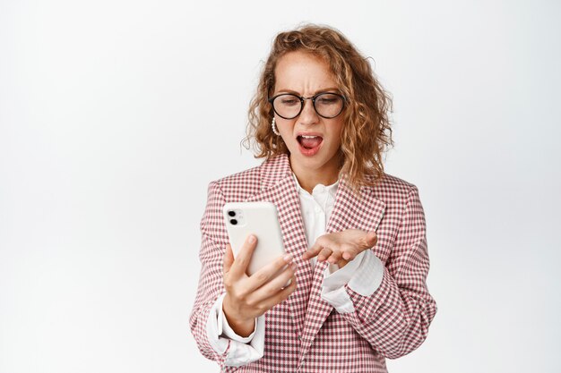 Mujer corporativa enojada, señora jefe mirando el teléfono móvil y gritando frustrado, parado blanco molesto.