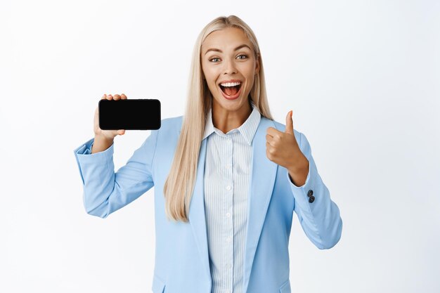 Mujer corporativa entusiasta que muestra la pantalla del teléfono móvil y los pulgares hacia arriba recomendando la aplicación de teléfono inteligente de pie en traje sobre fondo blanco