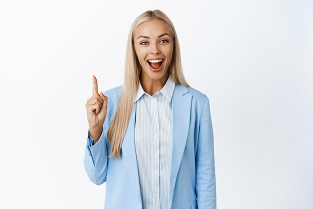 Mujer corporativa entusiasta señalando con el dedo sonriendo y mostrando un anuncio de pie en traje sobre fondo blanco.