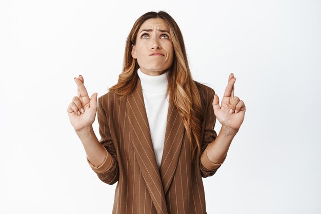 Mujer corporativa rezando con los dedos cruzados mirando hacia arriba suplicando esperanzados por un sueño hecho realidad haciendo un deseo de pie sobre fondo blanco