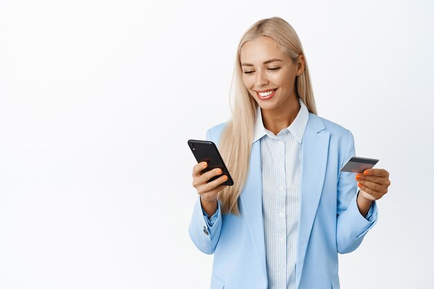 Mujer corporativa rubia sonriente ingrese información en el teléfono móvil con tarjeta de crédito con traje de pie sobre fondo blanco