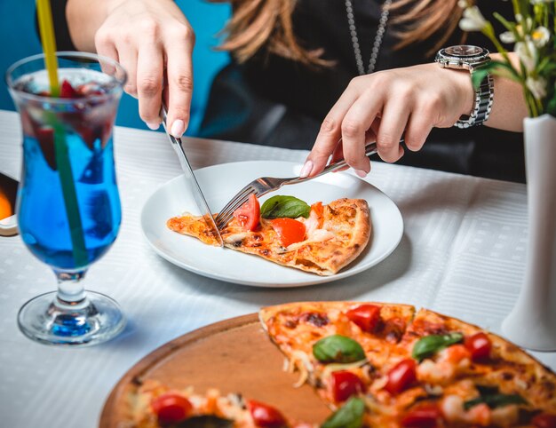 Mujer cortando una rebanada de pizza con cubiertos y un vaso de cóctel de laguna azul de pie alrededor.