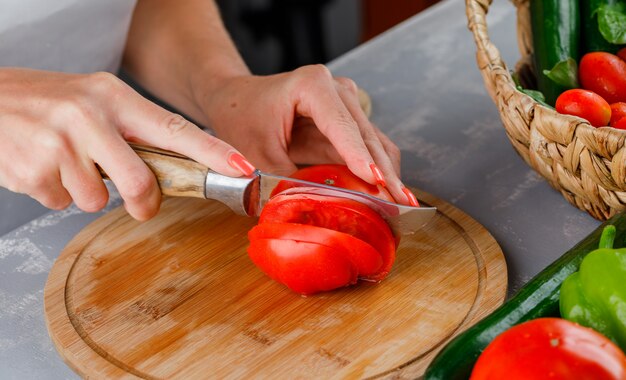 Mujer cortando tomate en una tabla de cortar vista de ángulo alto sobre una superficie gris