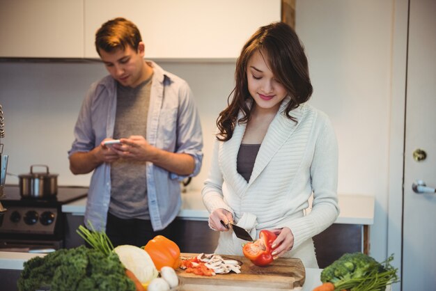 Mujer cortando verduras y hombre mediante teléfono móvil en la cocina