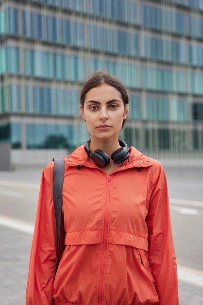 Mujer en cortavientos casual lleva karemat lleva auriculares estéreo alrededor del cuello entra para hacer deporte regularmente posa contra la construcción de la ciudad moderna