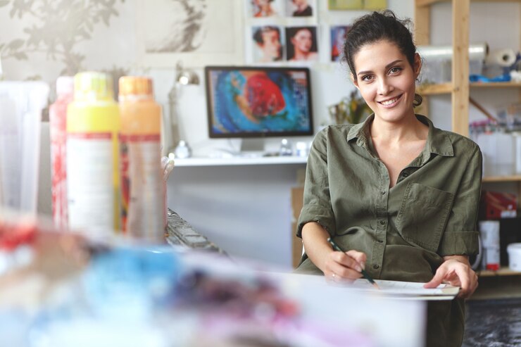 Foto gratuito mujer creativa joven positiva vestida casualmente, sentada en su taller, haciendo bocetos con lápiz, participando en el proceso creativo, disfrutando de su trabajo. concepto de personas, estilo de vida y arte