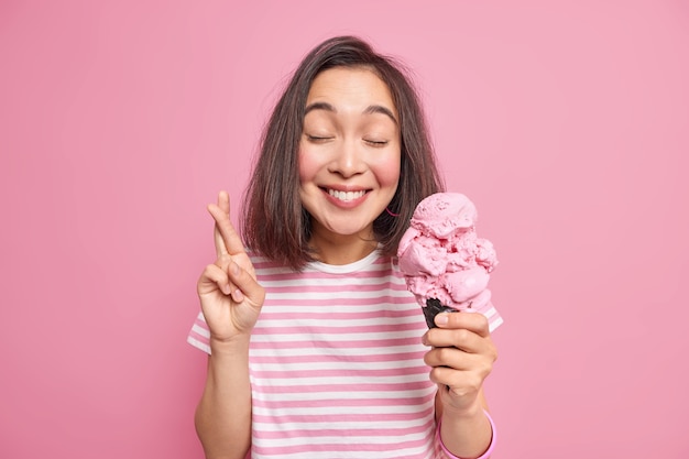 mujer cree en la buena fortuna mantiene los dedos cruzados cierra los ojos sonríe agradablemente viste una camiseta a rayas casual sostiene un apetitoso helado