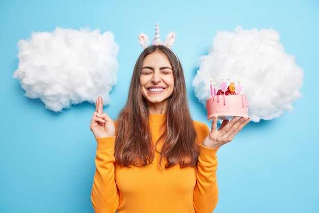 Mujer cruza los dedos hace deseo antes de quemar velas en la torta festiva sonríe ampliamente aislado en azul