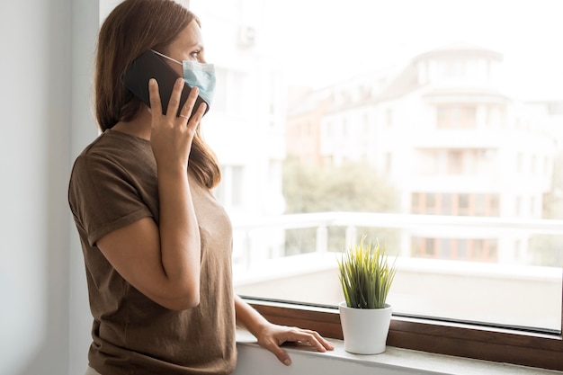 Mujer en cuarentena en casa hablando por teléfono mientras mira por la ventana