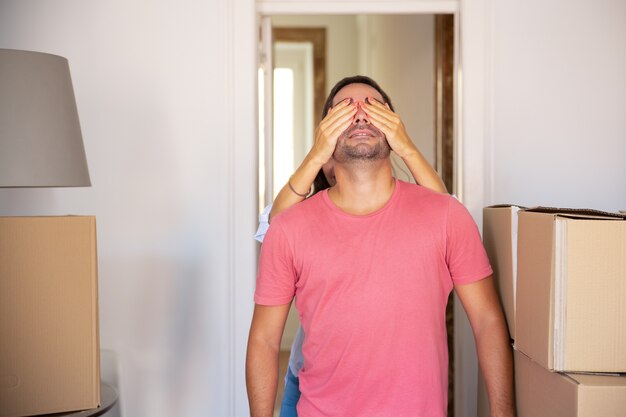 Mujer cubriendo los ojos de su novio con las manos y llevándolo a su nuevo apartamento con cajas de cartón