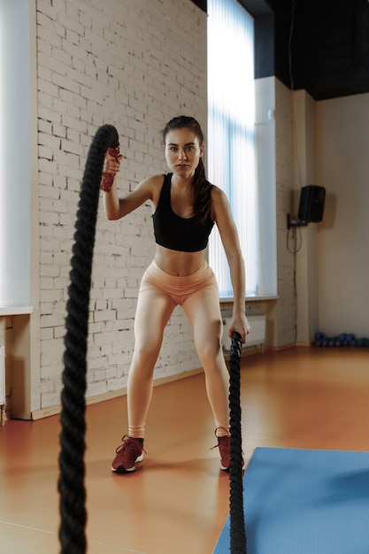 Mujer con cuerdas de batalla ejercicio en el gimnasio. Atleta, deporte, cuerda, entrenamiento, entrenamiento, ejercicios y concepto de estilo de vida saludable
