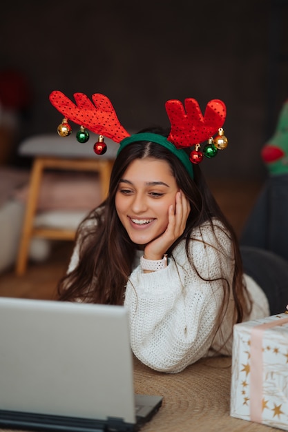 Mujer con cuernos sonriendo mientras habla con un amigo en línea en la computadora portátil durante la celebración de Navidad en casa