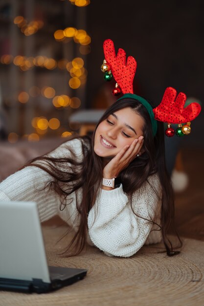 Mujer con cuernos sonriendo mientras habla con un amigo en línea en la computadora portátil durante la celebración de Navidad en casa