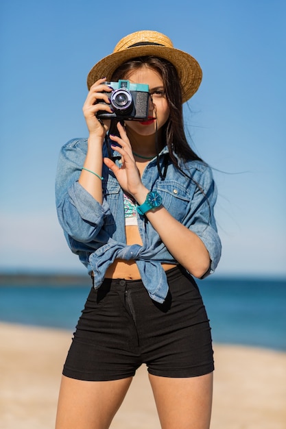 mujer con cuerpo bronceado, labios carnosos rojos y piernas largas posando en la playa tropical soleada. Vistiendo crop top, pantalones cortos y sombrero de paja.