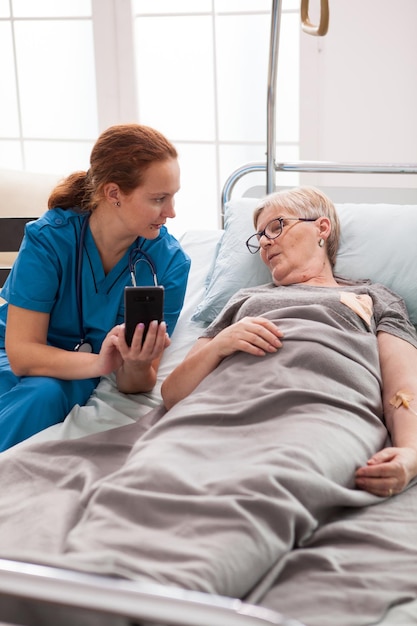 Mujer cuidadora en un hogar de ancianos hablando con una anciana sentada en la cama y usando un teléfono móvil.