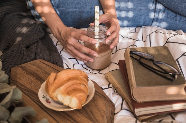 Mujer de cultivos con bebida cerca de libros y croissant