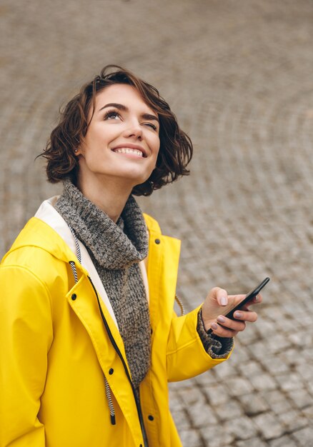 Mujer curiosa con cabello castaño rizado leyendo pronóstico en teléfono inteligente y mirando al cielo atornillando su ojo