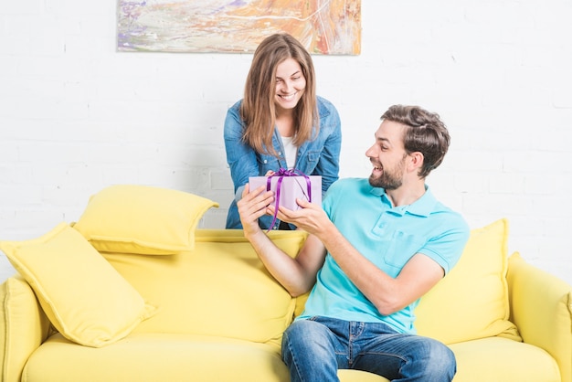 Mujer dando regalo sorpresa a su novio feliz sentado en el sofá