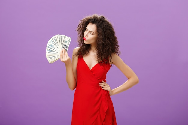 Mujer decidiendo cómo gastar mucho dinero en efectivo sosteniendo y mirando dinero doblando los labios tomando una decisión o t ...