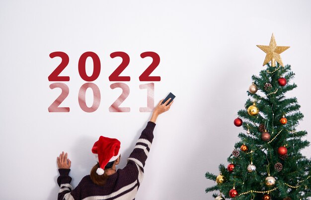Mujer decorando un árbol de Navidad y eliminando el texto 2021 en la pared en el día de año nuevo 2022.