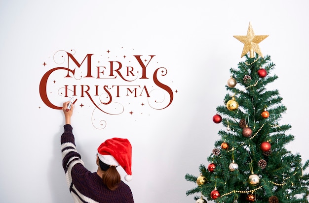 Mujer decorar un árbol de Navidad y escribir texto feliz Navidad en la pared en el día de Navidad