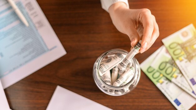 Mujer dejando caer un billete en un frasco con billetes enrollados sobre la mesa. Papeles, dinero en la mesa