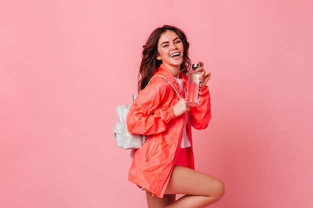 Mujer delgada activa en chaqueta deportiva y con mochila se ríe y sostiene una botella de agua en la pared rosa