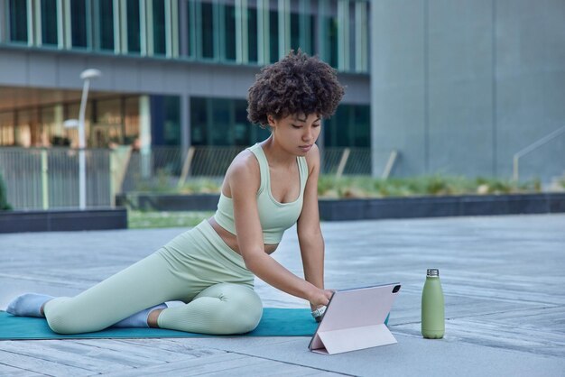 Mujer delgada activa practica yoga utiliza tableta digital para tener entrenamiento en línea relojes tutoriales bebe agua fresca vestida con ropa deportiva poses al aire libre contra el fondo borroso. Concepto de fitness