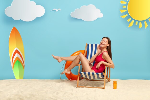 Mujer delgada y atractiva descansa en una tumbona en la playa, muestra piernas delgadas, usa bikini rojo, tiene un aspecto alegre