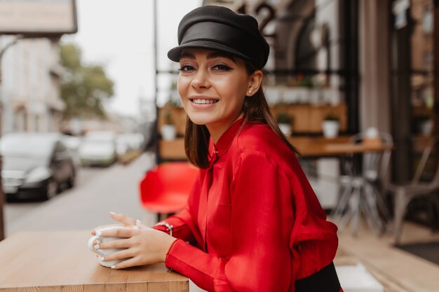 Mujer delgada con camisa de satén y sombrero mira suavemente a la lente. Señora de cabello castaño disfrutando de su bebida