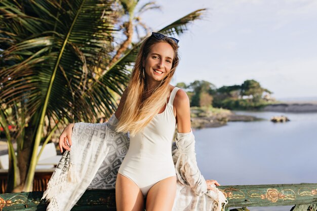 Mujer delgada disfrutando en un lugar exótico. Señora bronceada alegre de pie cerca de palmeras y sonriendo.