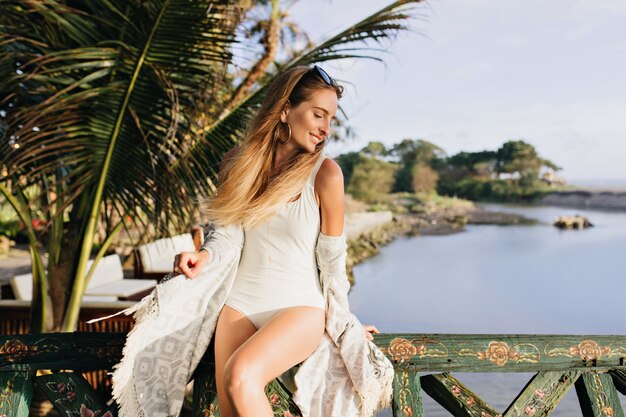 Mujer delgada disfrutando en un lugar exótico. Señora bronceada alegre de pie cerca de palmeras y sonriendo.