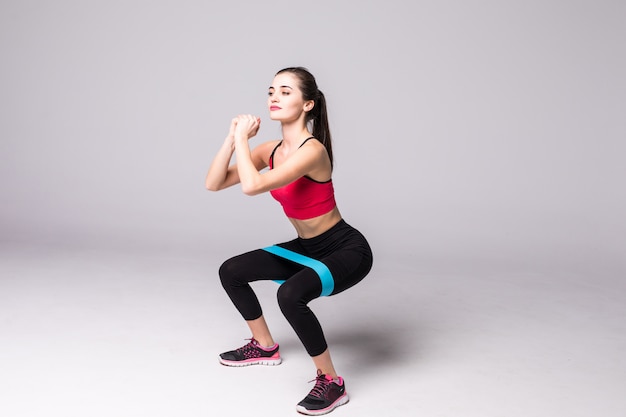 Mujer delgada haciendo sentadillas con banda de bucle elástico fitness aislada en la pared blanca