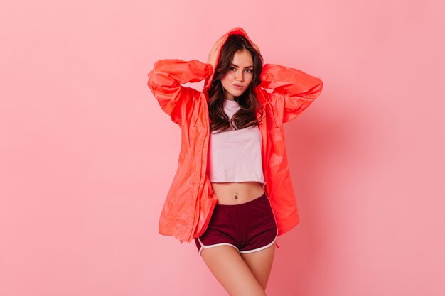 Mujer delgada en impermeable naranja posando en la pared rosa