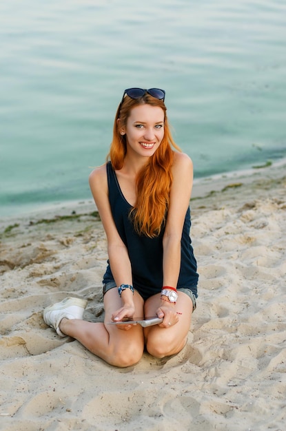 Mujer delgada pelirroja sonriente sentada en una playa y sosteniendo una tableta.