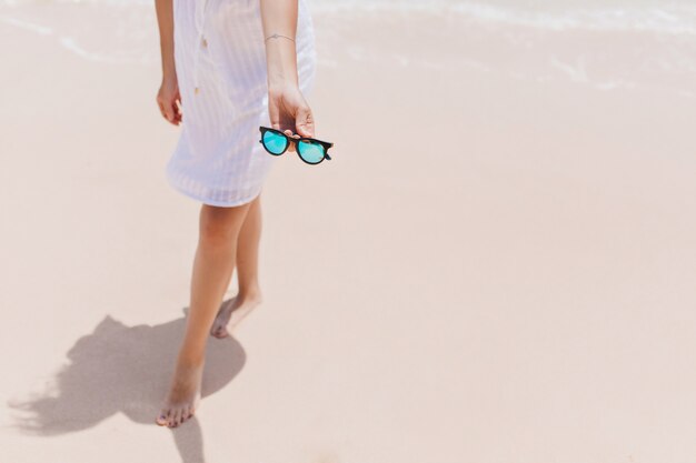 Mujer delgada posando en la costa con gafas de sol. Disparo al aire libre de relajada mujer descalza en vestido blanco de pie cerca del océano.