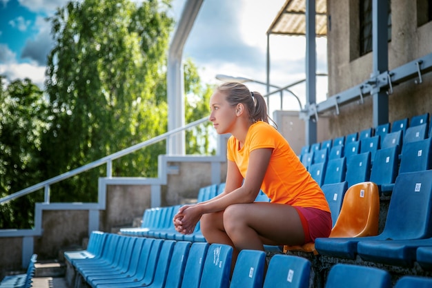 Mujer delgada con ropa deportiva naranja sentada en asientos de estadio de plástico azul.