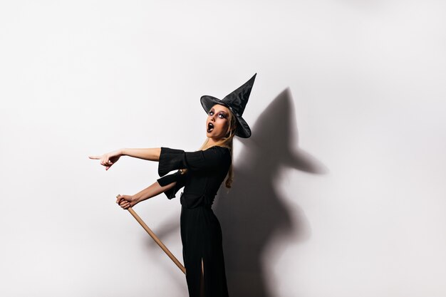 Mujer delgada sorprendida en traje de bruja posando. Filmación en interiores del fascinante mago con sombrero negro.