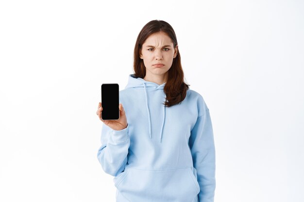 La mujer demostró la pantalla de su teléfono inteligente con cara triste, de pie contra la pared blanca