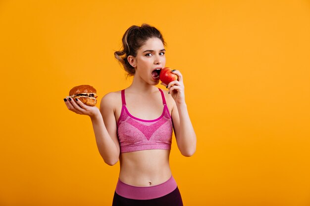 Mujer deportiva come manzana roja sobre pared naranja