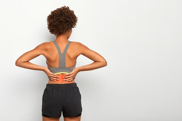 Mujer deportiva con corte de pelo afro toca la cintura con ambas manos, siente dolor en la columna vertebral, muestra la ubicación de la inflamación, usa top gris
