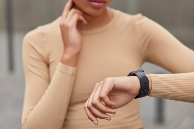 Mujer deportiva desconocida comprueba el pulso en el cuello centrado en smartwatch vestida con ropa deportiva beige plantea al aire libre