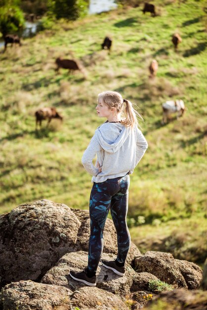 Mujer deportiva hermosa que se coloca en campo de roca con el fondo de las vacas.