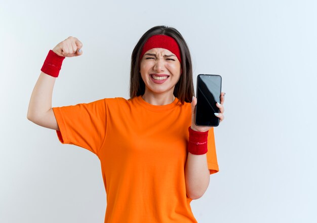 Mujer deportiva joven alegre con diadema y muñequeras que muestran el teléfono móvil haciendo un gesto fuerte aislado en la pared blanca con espacio de copia