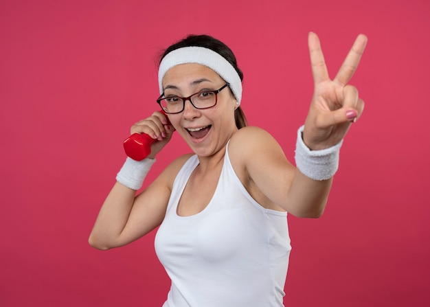 Mujer deportiva joven alegre en gafas ópticas con diadema y muñequeras sostiene mancuernas y gestos signo de mano de victoria aislado en la pared rosa