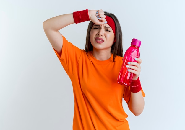 Foto gratuita mujer deportiva joven cansada con diadema y muñequeras sosteniendo una botella de agua tocando la frente aislada en la pared blanca con espacio de copia