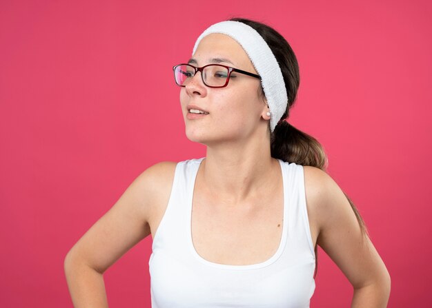 Mujer deportiva joven complacida en gafas ópticas con diadema y muñequeras mira al lado aislado en la pared rosa